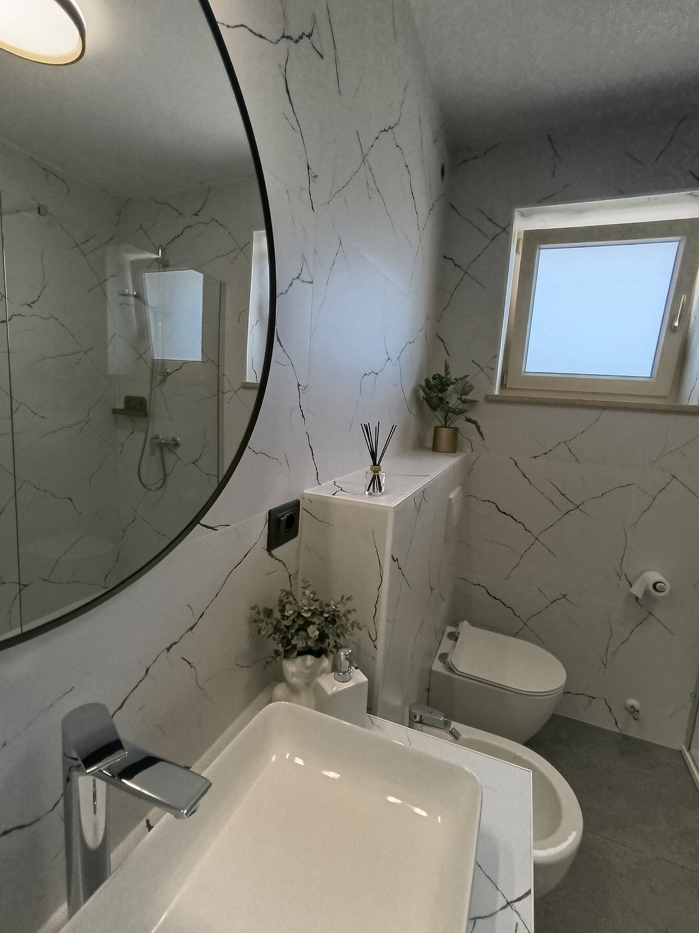 Une salle de bain avec un mur en marbre blanc, un grand miroir, un lavabo, des toilettes et une fenêtre.