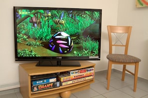 A television displaying a colorful animated scene, a wooden entertainment center with board games, and a wooden chair. - Central location, south-facing balcony, own ground-floor bicycle storage room, e-car charging station. (Binz)