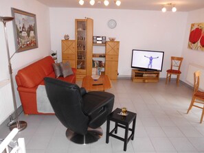 A living room with a red sofa, a black armchair, a wooden cabinet, a television, and a small table with a glass. - Central location, south-facing balcony, own ground-floor bicycle storage room, e-car charging station. (Binz)