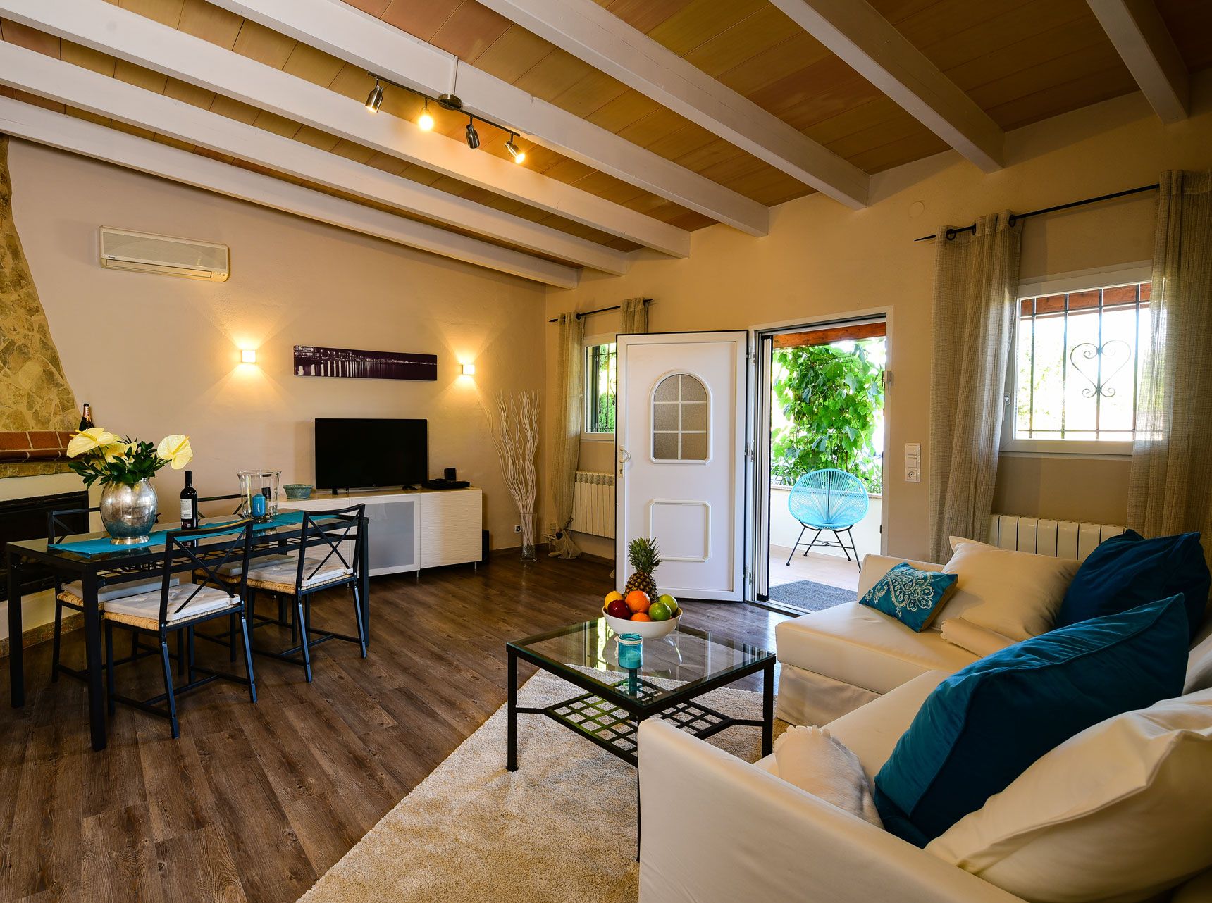 Un salon chaleureux avec un mur en pierre, des poutres apparentes en bois au plafond, un coin repas avec une table et des chaises, un canapé avec des coussins et une table basse en verre sur laquelle sont disposés des fruits.