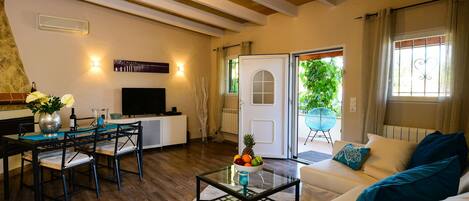 Un salon chaleureux avec un mur en pierre, des poutres apparentes en bois au plafond, un coin repas avec une table et des chaises, un canapé avec des coussins et une table basse en verre sur laquelle sont disposés des fruits.