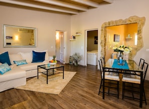 Un salon comprenant un canapé, une table basse et une table à manger avec des chaises. Un miroir est accroché au mur et une porte donne accès à une autre pièce.