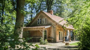 A rustic wooden cabin with a sloped roof, large windows, and a chimney, surrounded by dense trees. - historical house in Heidevillage on the outskirts of forest (Natendorf)