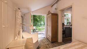 A bedroom with a bed, a bedside table, a chair, a wardrobe, and a view of the garden and kitchen through an open door. - historical house in Heidevillage on the outskirts of forest (Natendorf)