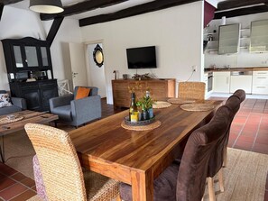 A dining area with a long wooden table, woven chairs, and a kitchen in the background.