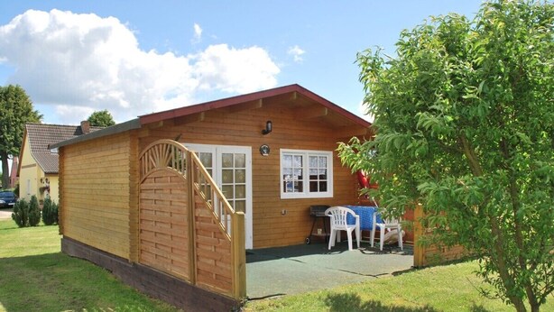 A wooden cabin with a wooden staircase leading to a door, a small patio with a table and chairs, and a tree in the foreground.