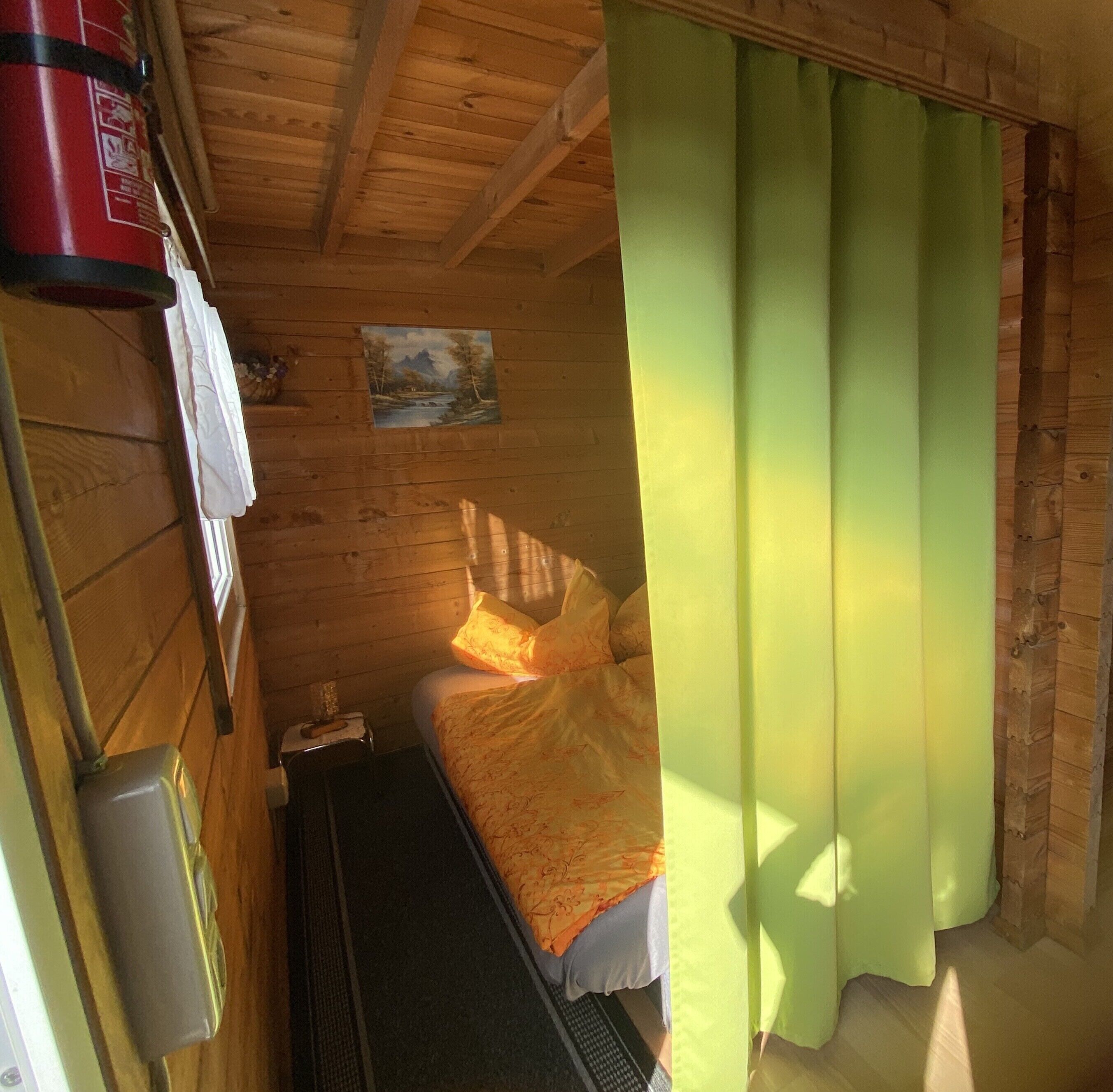 Une petite pièce chaleureuse aux murs en bois, avec un lit recouvert d’une couverture orange, un tableau accroché au mur et un rideau vert.