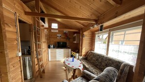 A wooden cabin interior with a sofa, a small table with a vase, a kitchen area, and a window with a view.