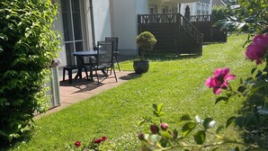 A residential building with a balcony, a small patio with a table and chairs, and a well-maintained lawn with flowers.