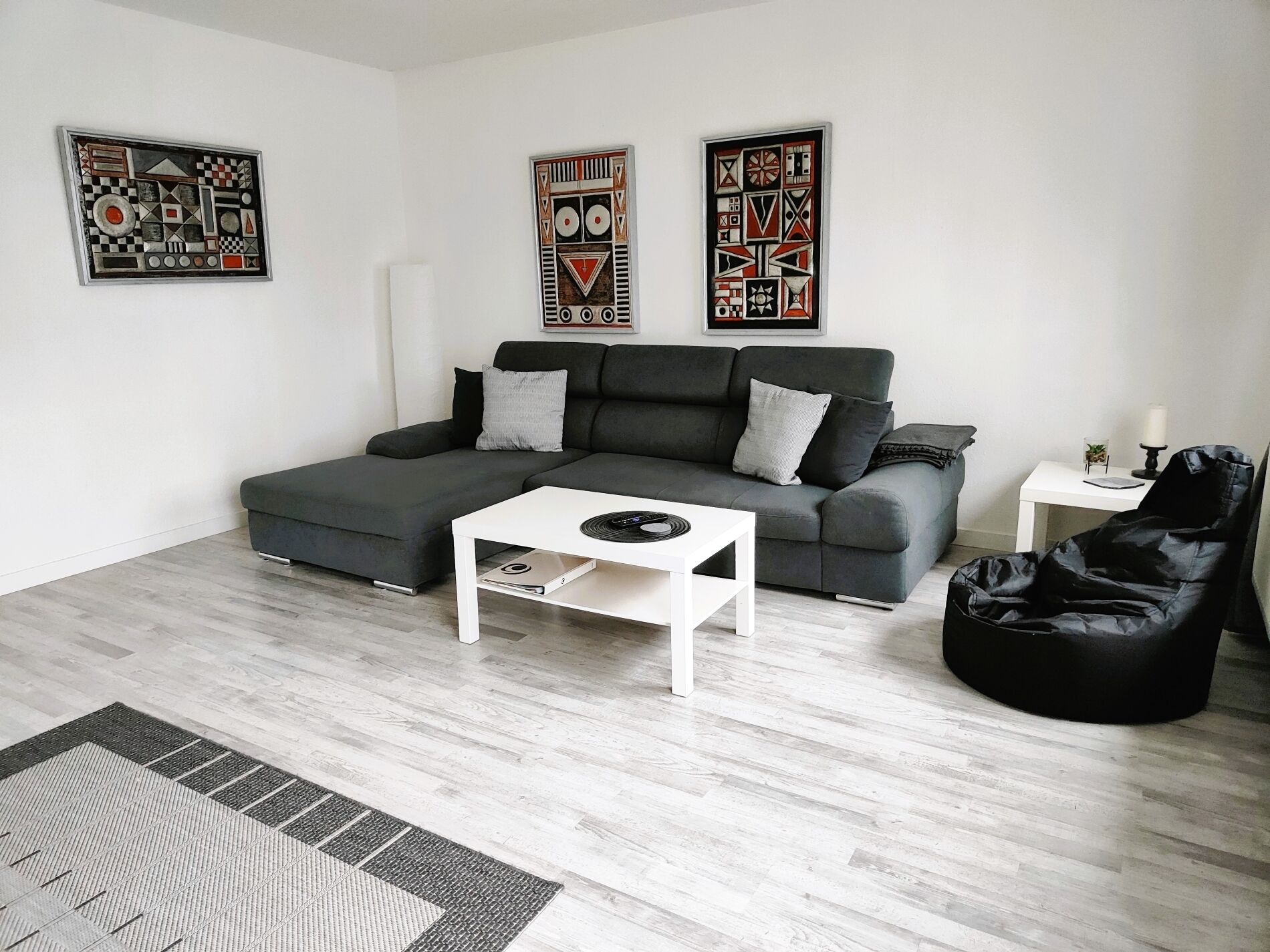 Uma sala de estar moderna com um sofá cinzento, uma mesa de centro branca, um pufe preto e três obras de arte abstratas nas paredes.