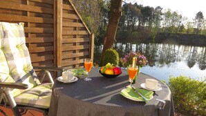 Une table dressée avec des aliments pour le petit-déjeuner, notamment des fruits et du jus, placée près d’une clôture en bois donnant sur une étendue d’eau.