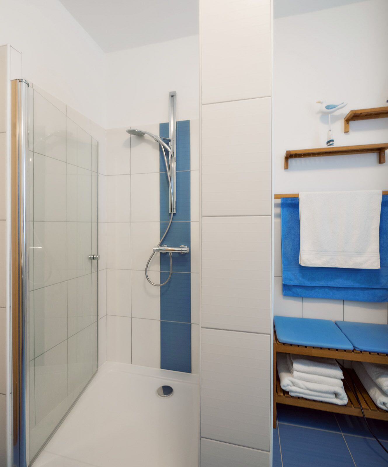 A modern bathroom with a glass-enclosed shower, a blue towel rack, and a wooden shelf with towels.