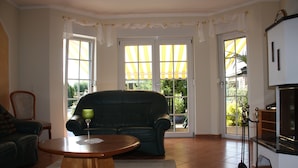 Un salon avec un canapé vert foncé, une table basse en bois et un vase en verre vert. On y voit deux fenêtres avec des rideaux, une porte et une partie de la cuisine.
