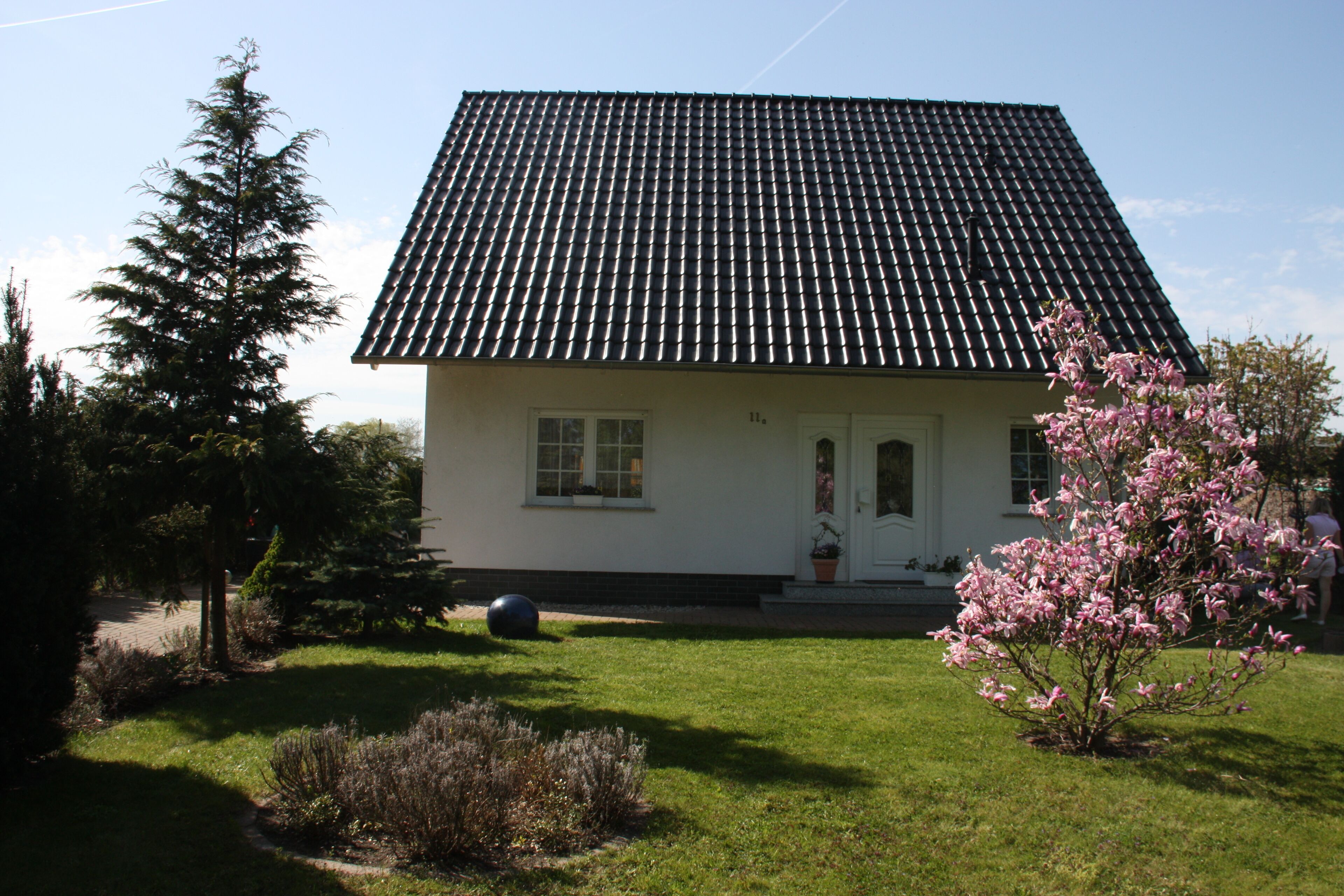 A house with a tiled roof, a front door, and a small garden with a tree and a flower bush.