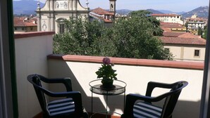 Ein Balkon mit zwei Stühlen und einem kleinen Tisch mit einem Blumentopf, mit Blick auf eine Stadt, in deren Hintergrund eine Kirche und Berge zu sehen sind.