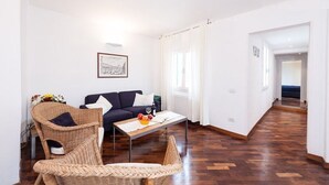 Ein Wohnzimmer mit einem dunkelblauen Sofa, Korbstühlen, einem Couchtisch mit Früchten und einem gerahmten Bild an der Wand.