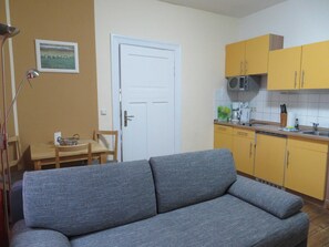 A living room with a sofa, a dining table with chairs, a white door, and a kitchen with yellow cabinets.