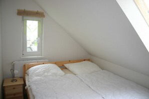 Ein kleines Zimmer mit einem Einzelbett, geneigter Decke, einem Fenster mit Jalousien, einem Nachttisch mit Lampe und einem Heizkörper.