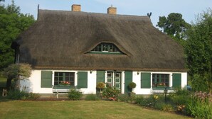 Ein traditionelles Haus mit Strohdach, grünen Fensterläden, einem kleinen Garten mit Blumen und blauem Himmel.