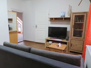 Un salon comprenant un canapé, un téléviseur posé sur un meuble en bois, un buffet aux portes vitrées et une porte donnant sur une autre pièce.