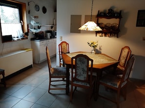 Ruokailutila, jossa on puinen pöytä ja tuolit, riippuvalaisin sekä seinälle kiinnitetty hylly, jossa on koriste-esineitä.