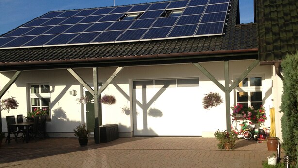Un bâtiment doté d’un toit recouvert de panneaux solaires, d’un patio abrité avec des sièges extérieurs, et d’un chemin en briques menant à l’entrée.