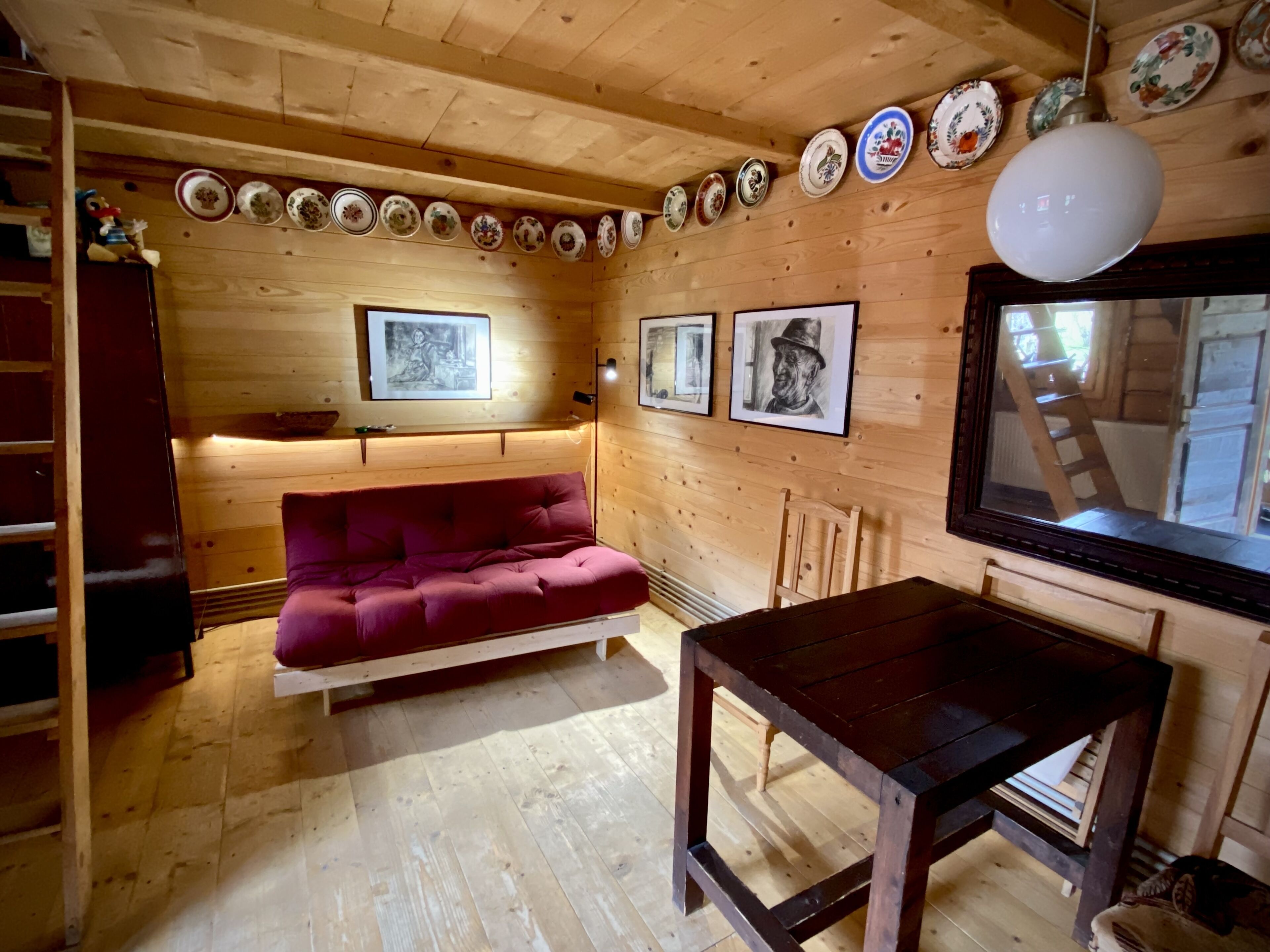 A cozy cabin interior with a wooden floor, a red sofa, a wooden table, and a row of decorative plates on the wall.