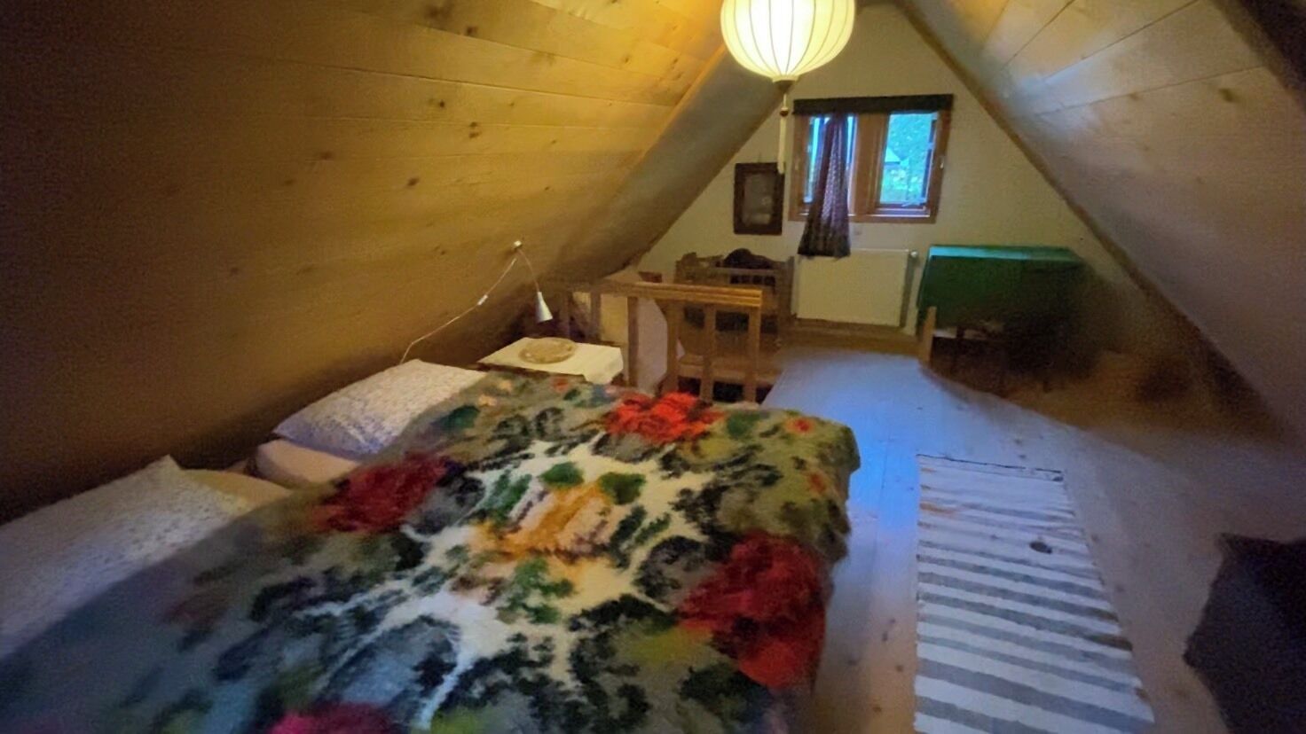 A small, cozy attic bedroom with a sloped ceiling, a bed with a colorful blanket, a desk with a chair, and a window with curtains.