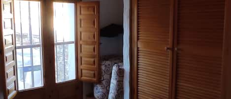 Una habitación con cama, una ventana con contraventanas abiertas, un armario de madera y un banco con cojines.