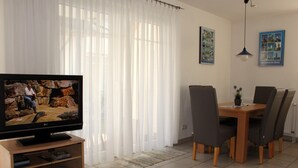 Ein Wohnzimmer mit Fernseher, einem Esstisch mit Stühlen, einem Bücherregal und einem Fenster mit durchsichtigen Vorhängen.