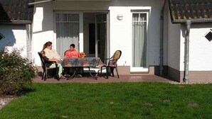 Eine Terrasse mit Tisch und Stühlen, ein überdachter Bereich und eine Rasenfläche vor einem Gebäude mit einer Tür und Fenstern.