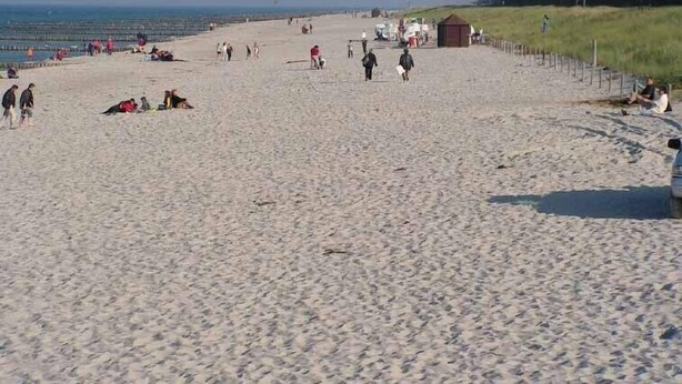 Ein weiter Sandstrand mit Menschen, die spazieren gehen und sitzen, einer kleinen Bebauung und einem grasbewachsen Hügel im Hintergrund.
