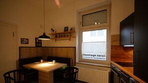 Une cuisine avec une table en bois, deux chaises, un luminaire suspendu et une fenêtre avec des stores.