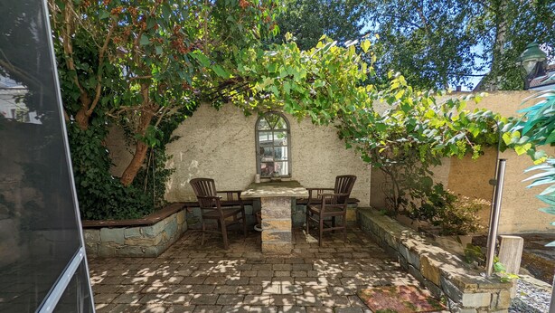 Une terrasse avec un sol en pierre, une table et des chaises, entourée de verdure et d’un mur en pierre.