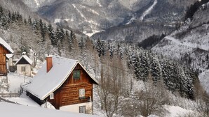 Una baita di legno con tetto imbiancato dalla neve, circondata da alberi e montagne innevate sullo sfondo.
