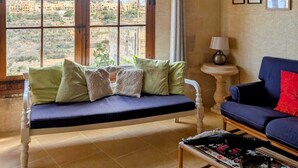 Una habitación acogedora con un amplio ventanal de arco que ofrece vistas a edificios y vegetación. Hay un sofá azul con cojines, una mesita con una lámpara y una bandeja de mimbre.
