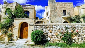 Un edificio de piedra tradicional con puerta de madera, ventanas arqueadas y un muro de piedra con vegetación.