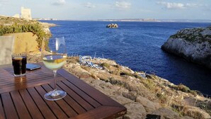 Una mesa con una copa de vino blanco y una copa de bebida oscura, con vistas a la costa, terreno rocoso y una isla distante.