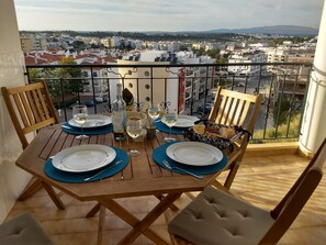 A balcony table set with plates, glasses, and a bottle, overlooking a cityscape with buildings and a distant mountain.