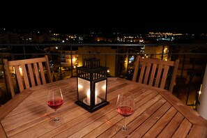 Et træbord med to vinglas og en lygte på en balkon med udsigt over en by natten.