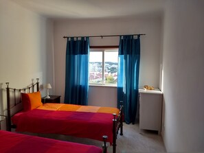 A room with two beds, a window with curtains, a nightstand, and a radiator.