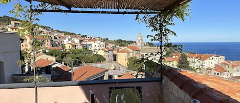 A terrace with a table, a bottle, and two glasses, overlooking a coastal town with buildings and a church.