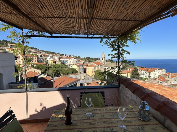 A terrace with a table, a bottle, and two glasses, overlooking a coastal town with buildings and a church. - Holiday house with sea view and sun terrace, 4 km to beach (Cipressa)