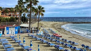 A beach with rows of blue lounge chairs and umbrellas, palm trees, and a view of the ocean. - Holiday house with sea view and sun terrace, 4 km to beach (Cipressa)