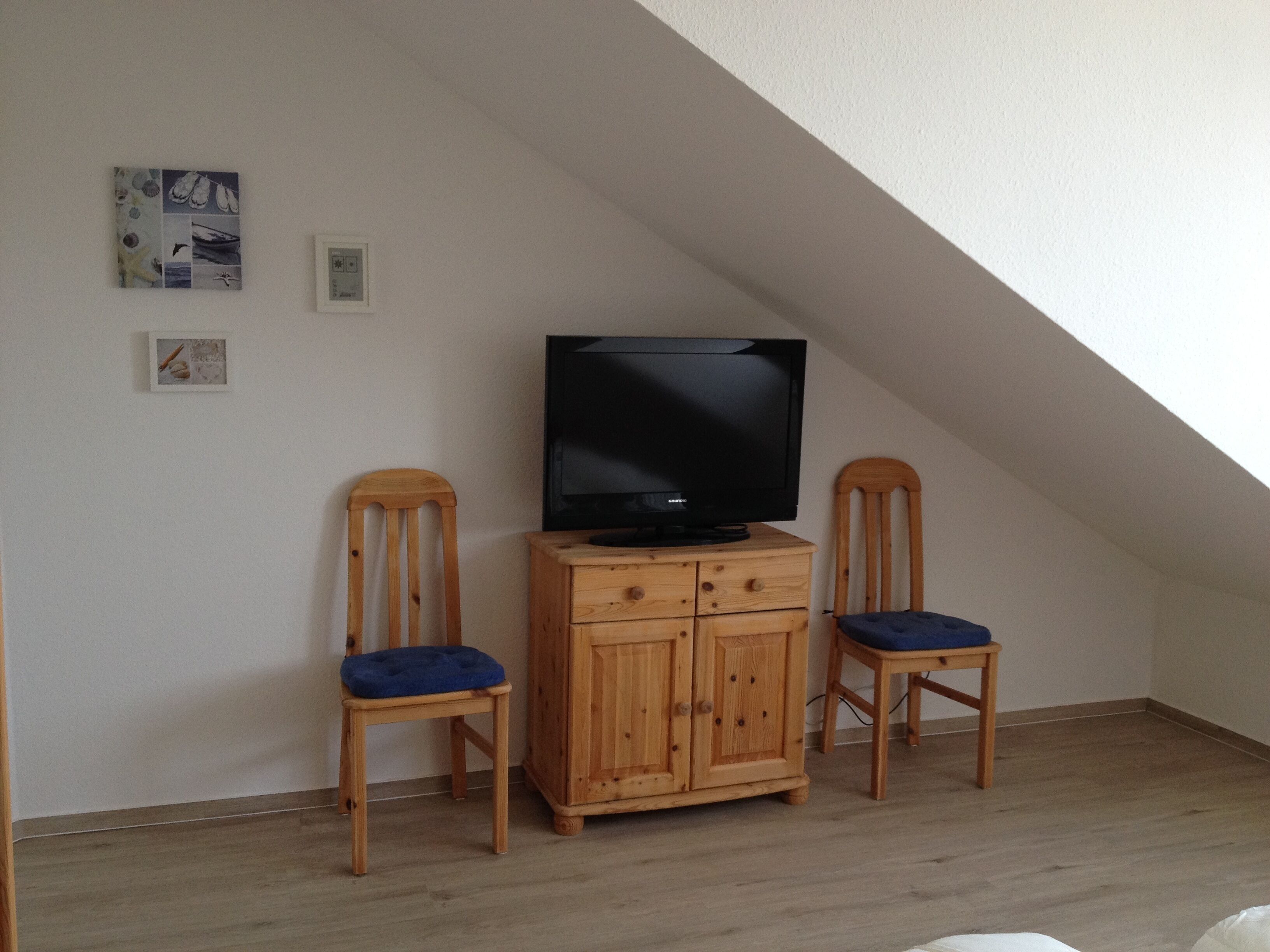 A room with a sloped ceiling, a wooden cabinet with a television on top, two wooden chairs with blue cushions, and a small wall-mounted shelf with framed pictures.