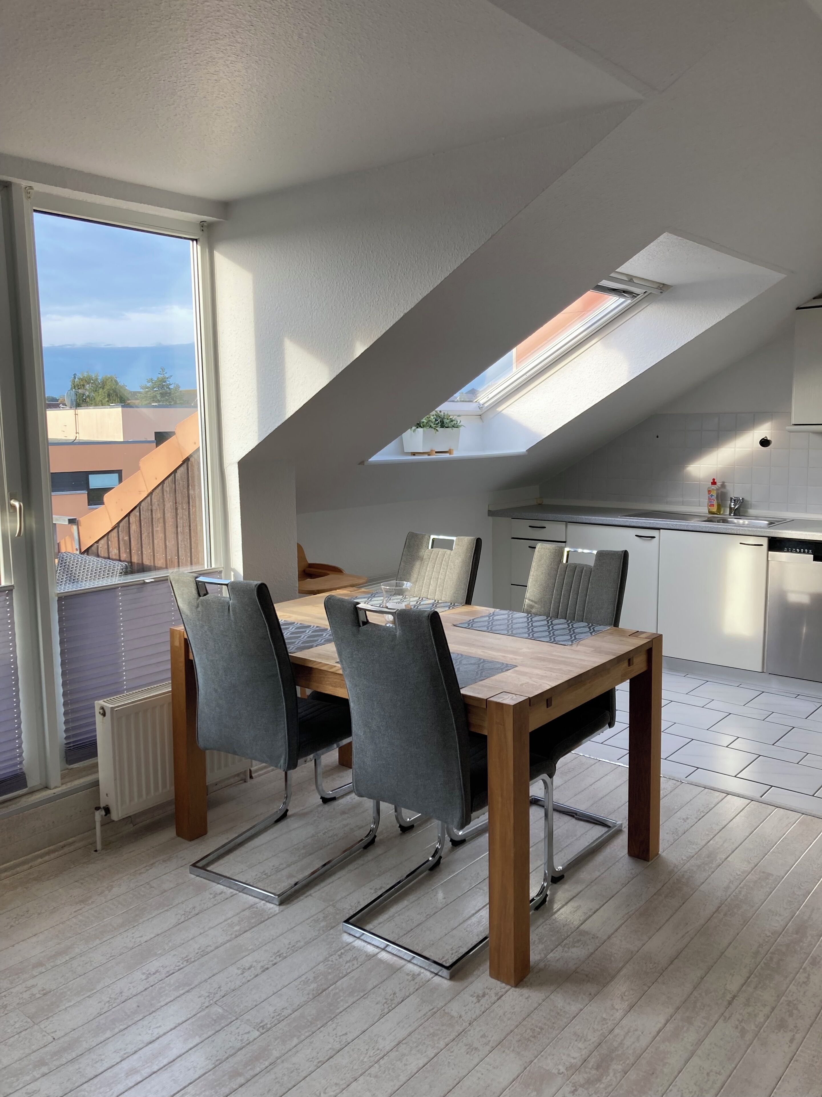 A modern kitchen with white cabinets, a dining area with a wooden table and grey chairs, and a skylight above the dining area.