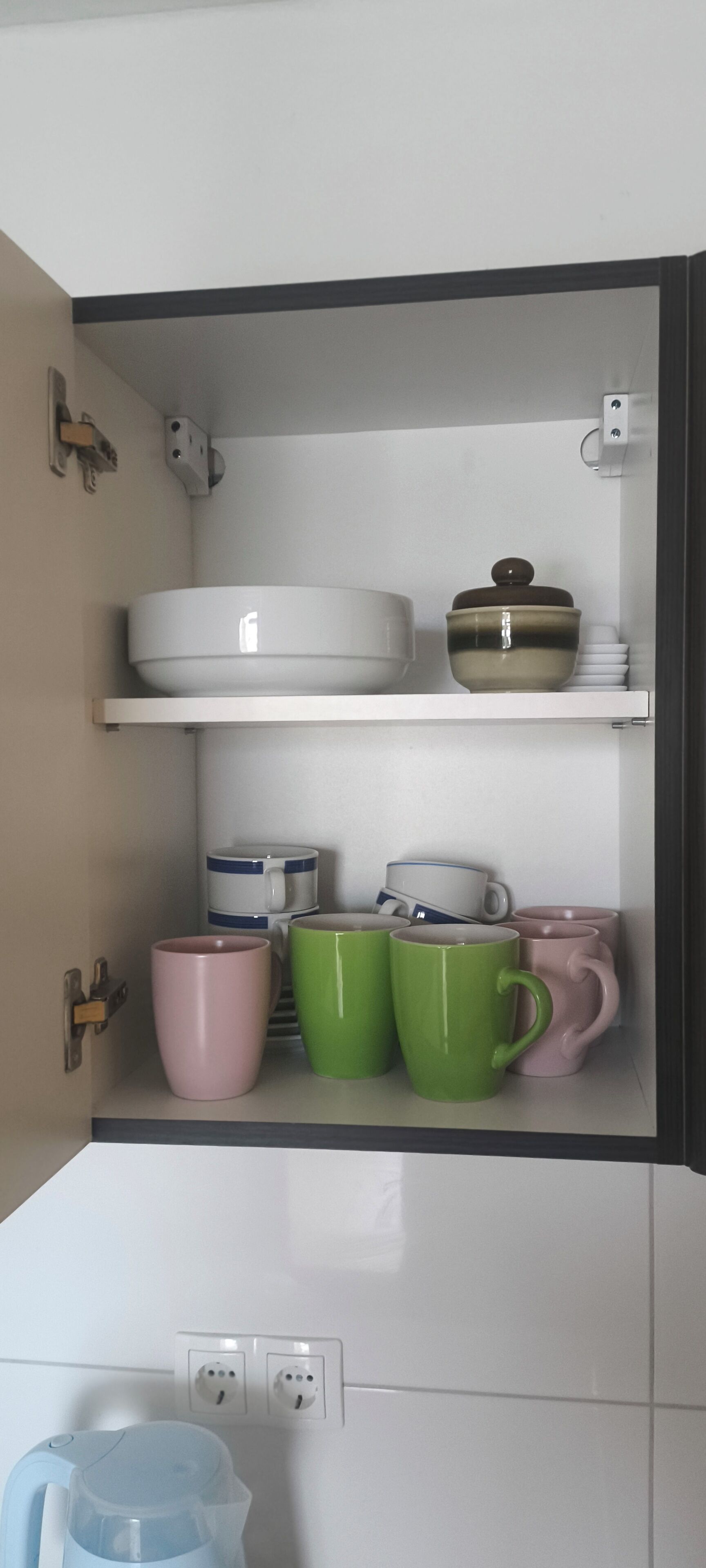 A kitchen cabinet with white and green mugs, a white bowl, and a brown pot.