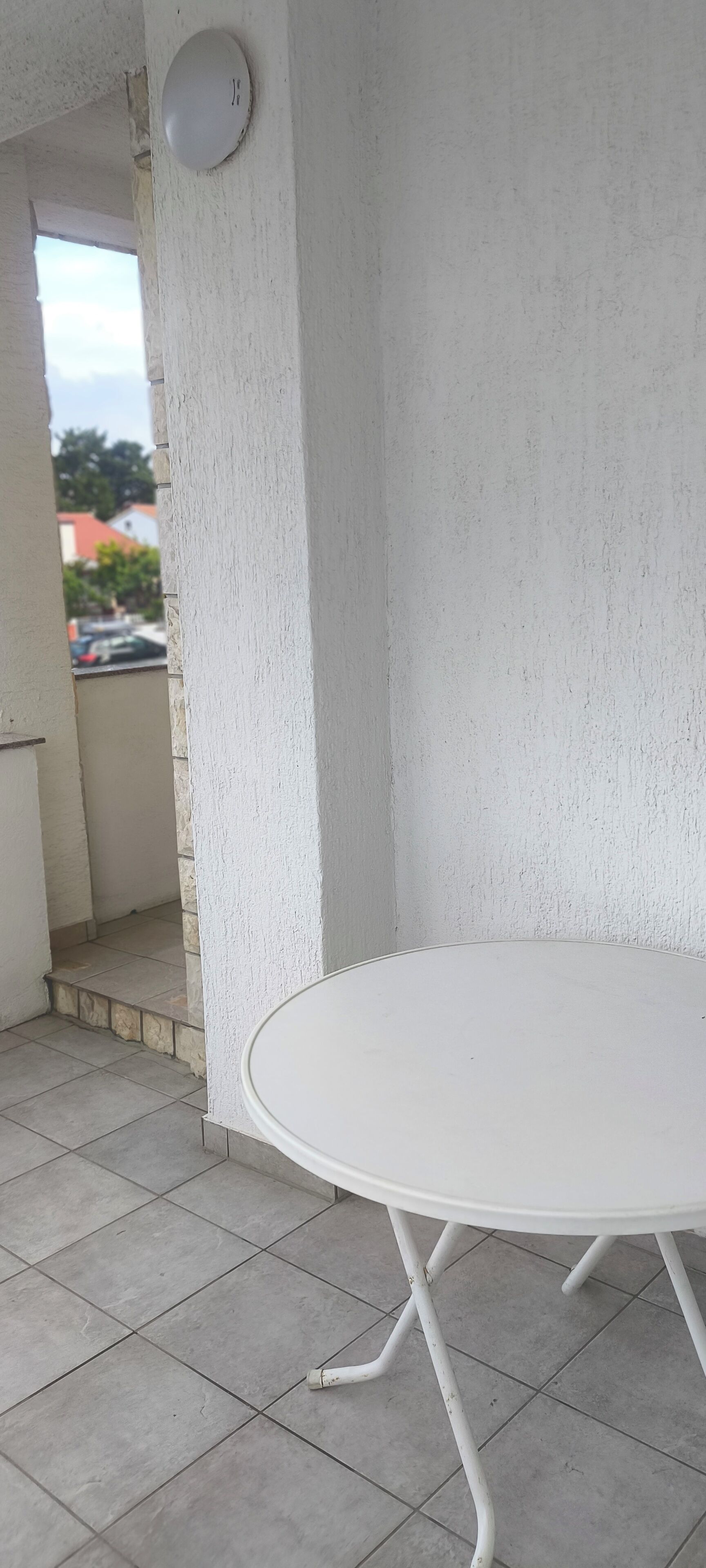Une table blanche aux pieds métalliques, posée sur un sol carrelé, un mur blanc et une fenêtre visible donnant sur des immeubles et des arbres.