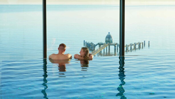 Un couple se trouve dans une grande piscine à eau claire, d’où l’on aperçoit une jetée au loin et une structure en forme de dôme.