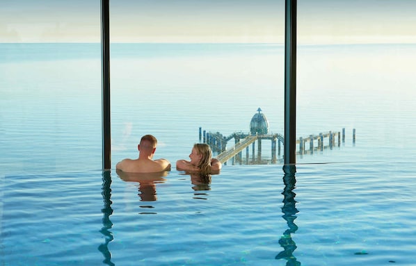 Um casal em uma piscina ampla e transparente, com vista para um cais distante e uma estrutura em forma de cúpula.